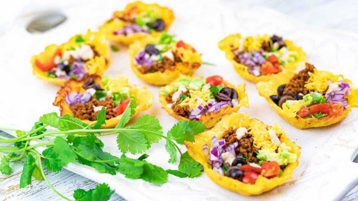 Mini taco cups filled with seasoned ground meat, shredded cheese, lettuce, tomatoes, olives, and cabbage deliver savory flavors in every bite, served on a white tray with fresh cilantro garnish.