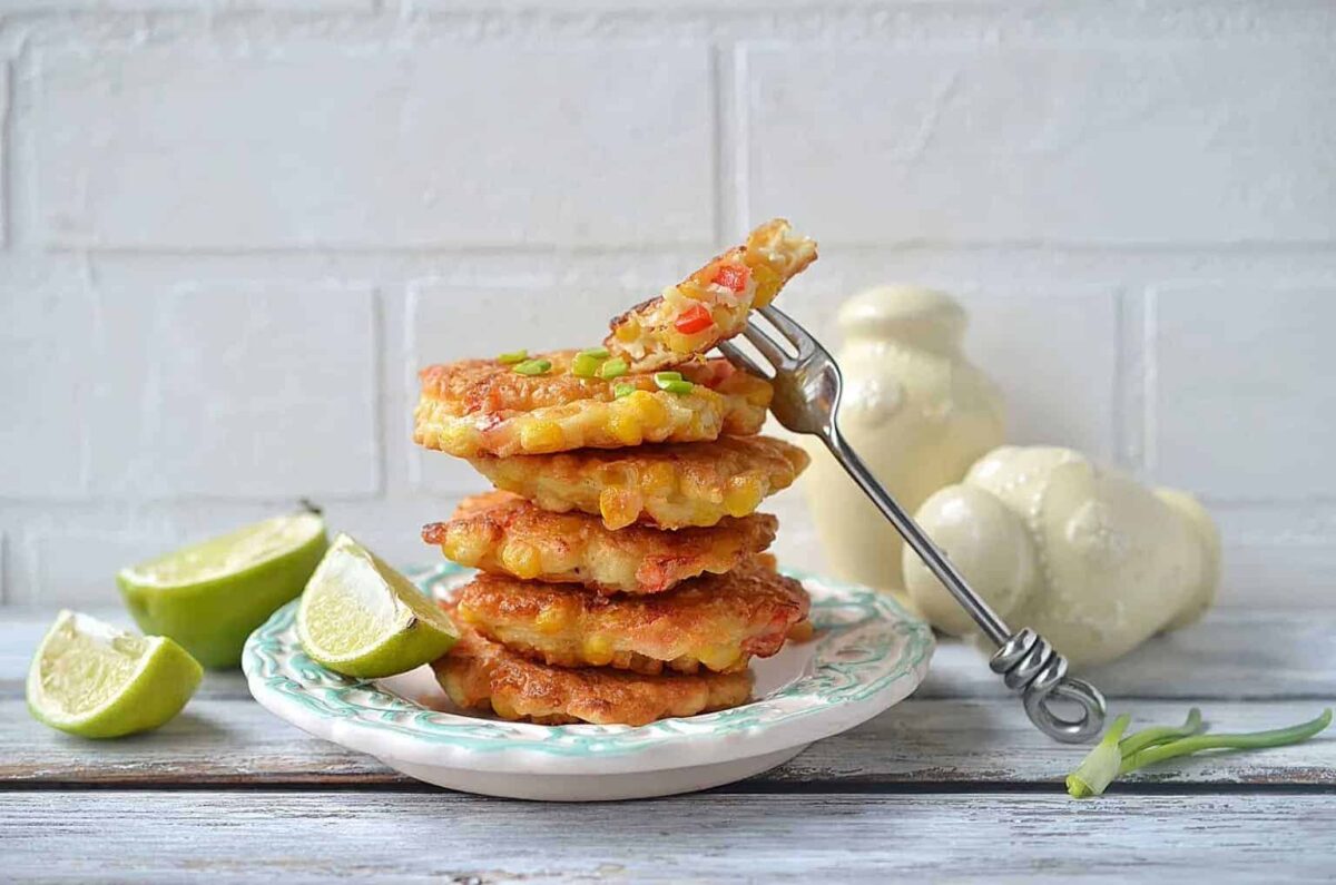 A stack of corn fritters on a plate with lime wedges, garnished with green onions, and a fork holding a piece—perfect for quick sides that add flavor to any meal.