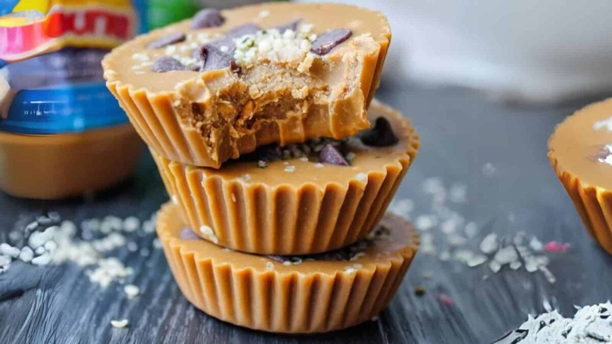 Three stacked peanut butter cups, the top one with a bite taken out, topped with chocolate chips and seeds, showcase sweet flavors and creative cup recipes on a dark surface.