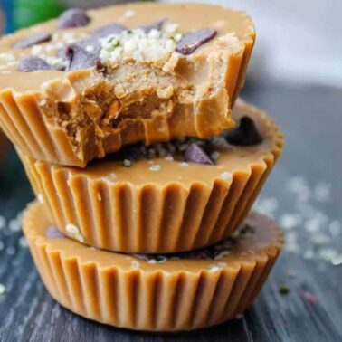 Three stacked peanut butter cups, the top one with a bite taken out, topped with chocolate chips and seeds, showcase sweet flavors and creative cup recipes on a dark surface.