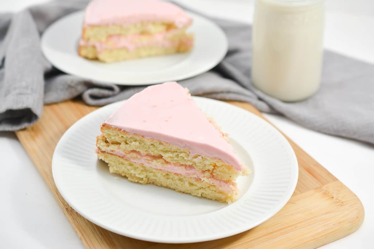 A slice of layered vanilla cake with pink frosting on a white plate, set on a wooden board with a glass of milk and a gray napkin in the background, highlights the comforting flavor found in classic vanilla recipes.