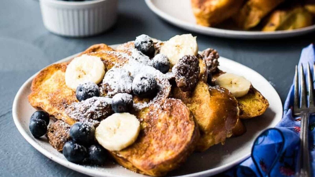 A plate of comforting French toast topped with sliced bananas, blueberries, chocolate pieces, and powdered sugar, offering a sweet flavor inspired by classic vanilla recipes. A fork and napkin sit beside the plate.