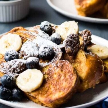 A plate of comforting French toast topped with sliced bananas, blueberries, chocolate pieces, and powdered sugar, offering a sweet flavor inspired by classic vanilla recipes. A fork and napkin sit beside the plate.