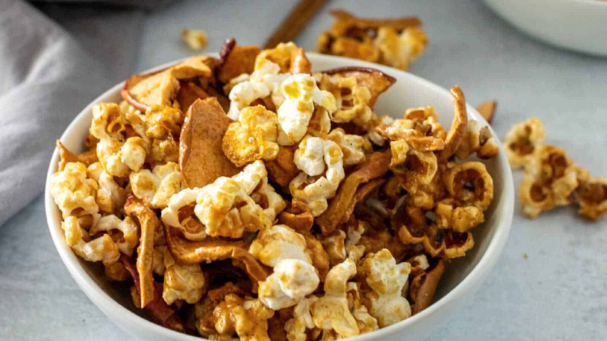 A white bowl filled with popcorn mixed with dried apple slices sits on a light surface, with some pieces scattered around it—perfect for easy snacks and creative snack ideas.