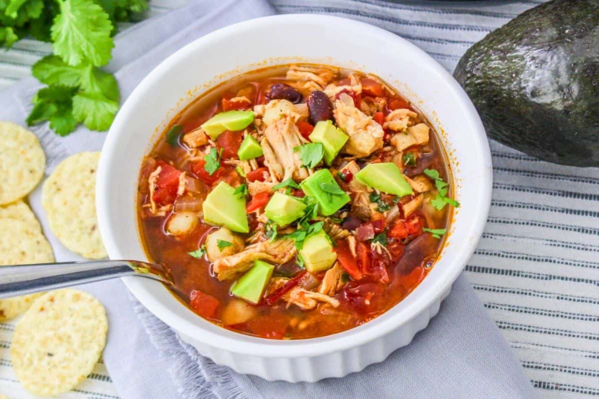A bowl of chicken chili soup topped with diced avocado and cilantro, with a spoon inside. Surrounded by tortilla chips, cilantro, and an avocado on a striped cloth—bringing together favorite flavors just like your own kitchen or favorite copycat dishes.