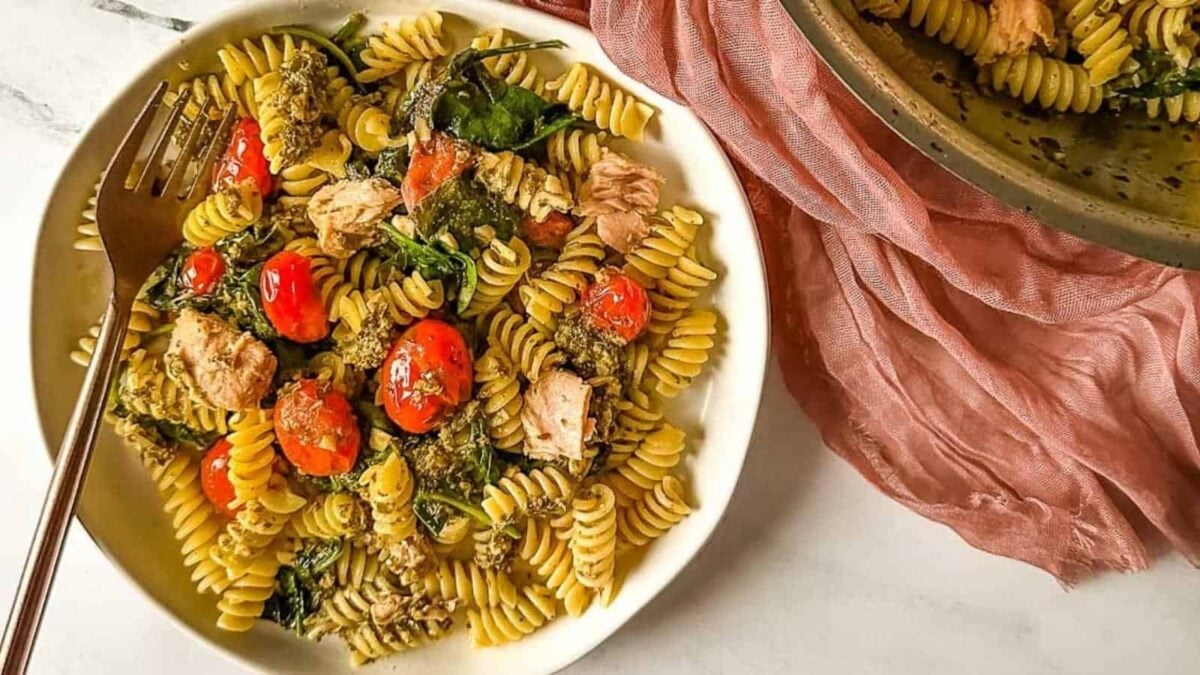 A bowl of rotini pasta with savory chunks of tuna, cherry tomatoes, spinach, and mushrooms, with a fork on the side and a pink cloth partially visible beside the bowl.