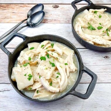 Two black bowls filled with chicken and dumplings, topped with chopped parsley, are set on a wooden surface next to two metal spoonsโa comforting copycat recipe inspired by restaurant favorites.