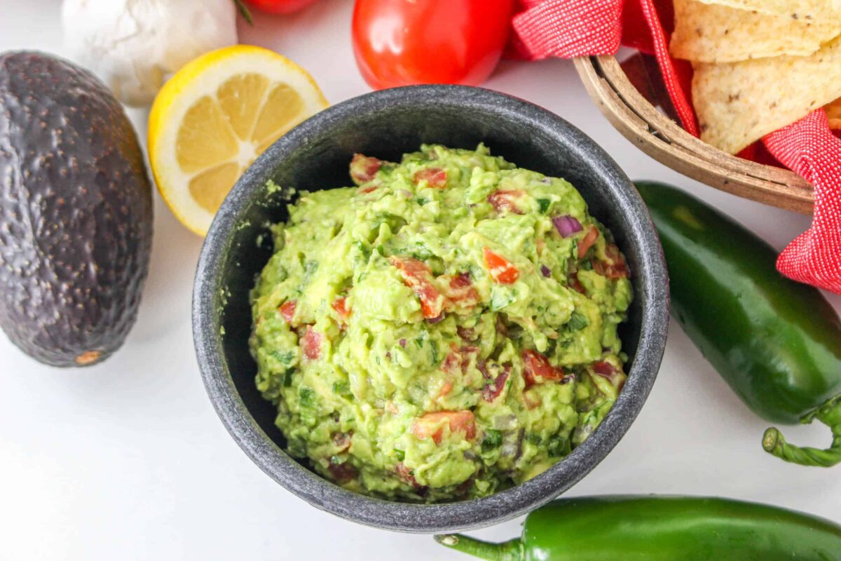 A bowl of guacamole with visible tomato and onion pieces, surrounded by avocado, lemon, jalapeños, tomatoes, garlic, and tortilla chips—perfect for enjoying fresh flavors in your favorite spring recipes.