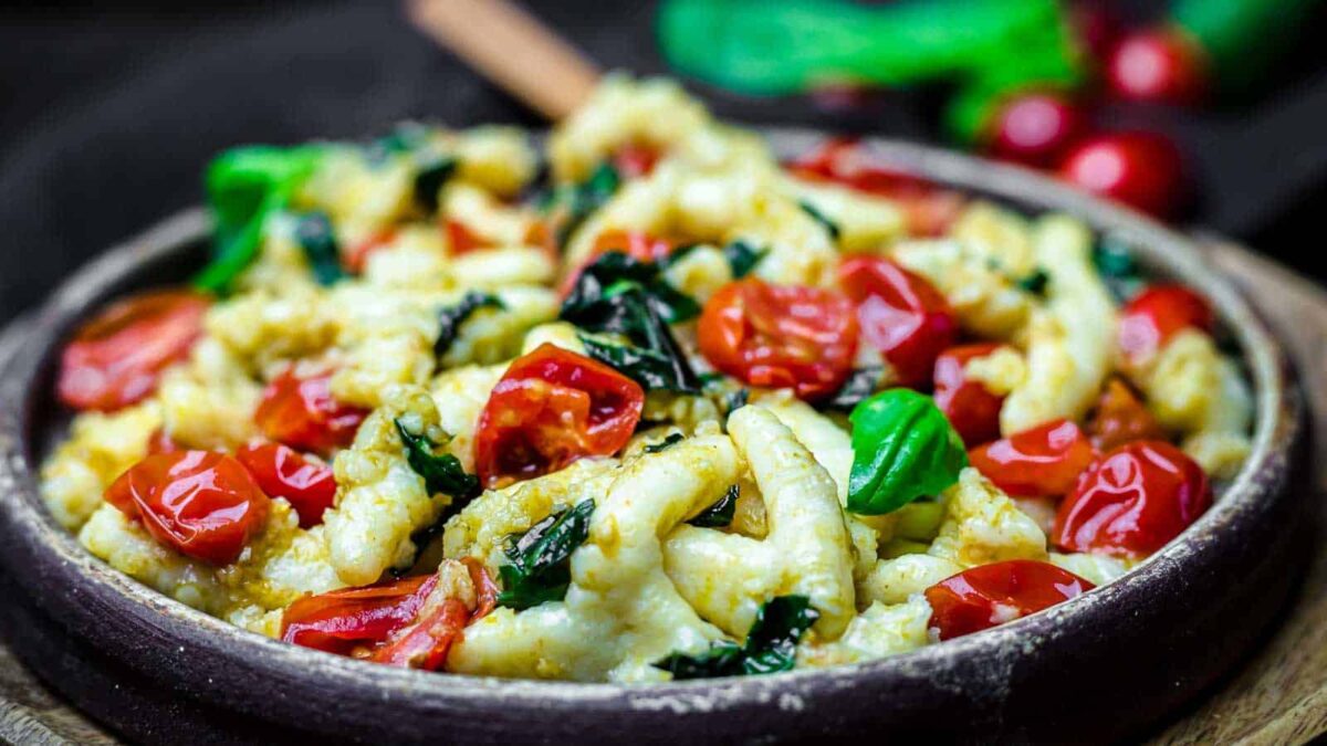 A bowl of pasta topped with wilted spinach, cherry tomatoes, and fresh basil leaves, capturing the fresh flavors of spring recipes and served in a rustic wooden bowl.