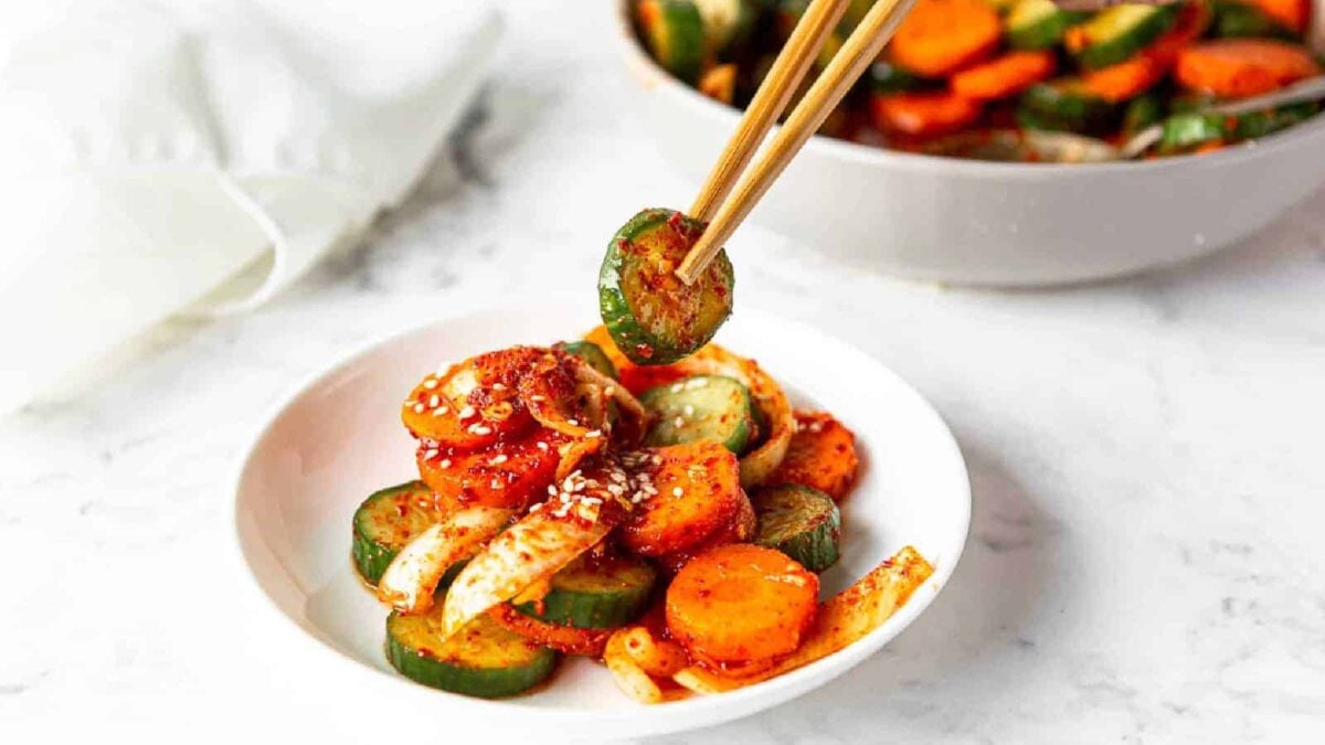 A bowl of sliced cucumber, carrot, and onion salad with sesame seeds and red seasoning delivers a cool crunch and fresh flavor, as chopsticks hold a cucumber slice above the bowl—a refreshing idea for easy cucumber recipes.