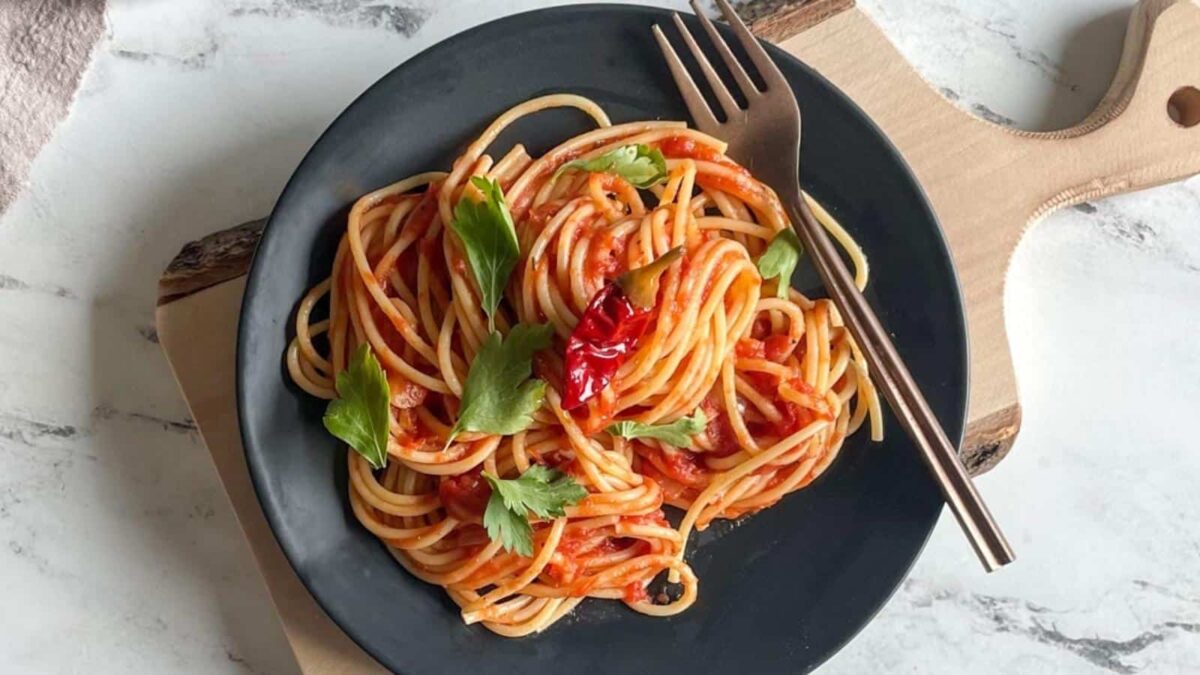 A black plate of spaghetti with hearty sauce, garnished with fresh herbs and a red chili pepper, sits on a wooden board with a fork resting on the plate—perfect for fans of classic Italian recipes.