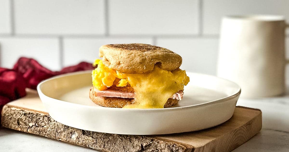 An egg sandwich with savory sandwich fillings—scrambled eggs, melted cheese, and ham—sits stacked between an English muffin on a white plate and wooden board.