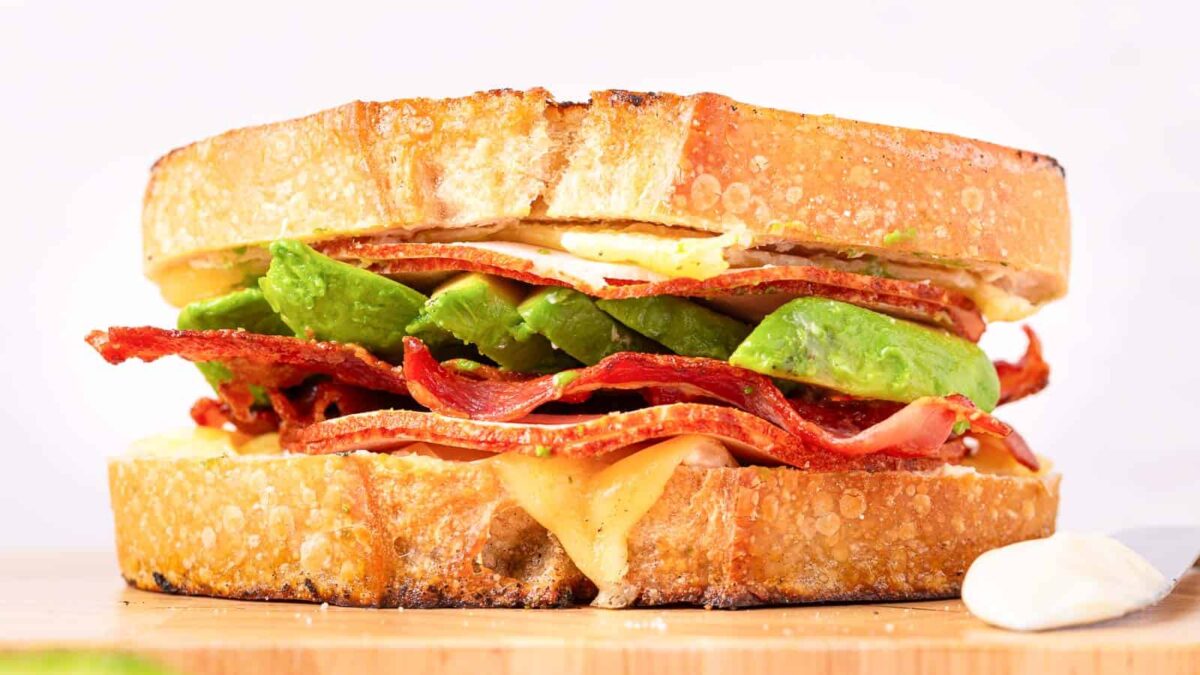 A close-up of a sandwich with stacked layers of thick toasted bread, melted cheese, bacon strips, avocado slices, and savory fillings, finished with sauce on a wooden board.