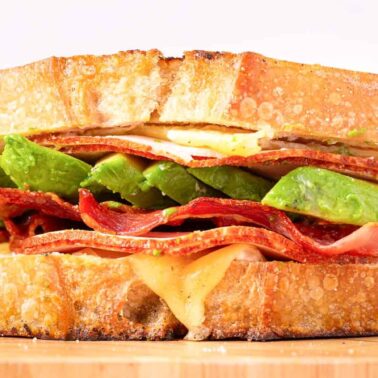 A close-up of a sandwich with stacked layers of thick toasted bread, melted cheese, bacon strips, avocado slices, and savory fillings, finished with sauce on a wooden board.