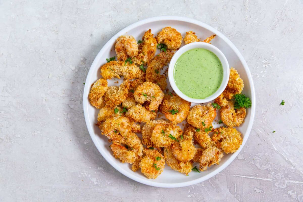 A white plate with breaded, baked shrimp garnished with parsley, served with a small bowl of green dipping sauce—perfect as party appetizers or crispy appetizers made easily in the air fryer.