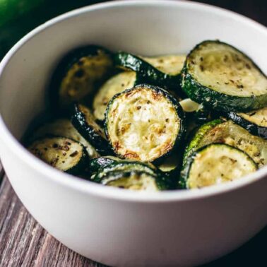 A white bowl filled with sliced, roasted zucchini, perfect for serving as crispy appetizers or new air fryer appetizer ideas, placed on a wooden surface.