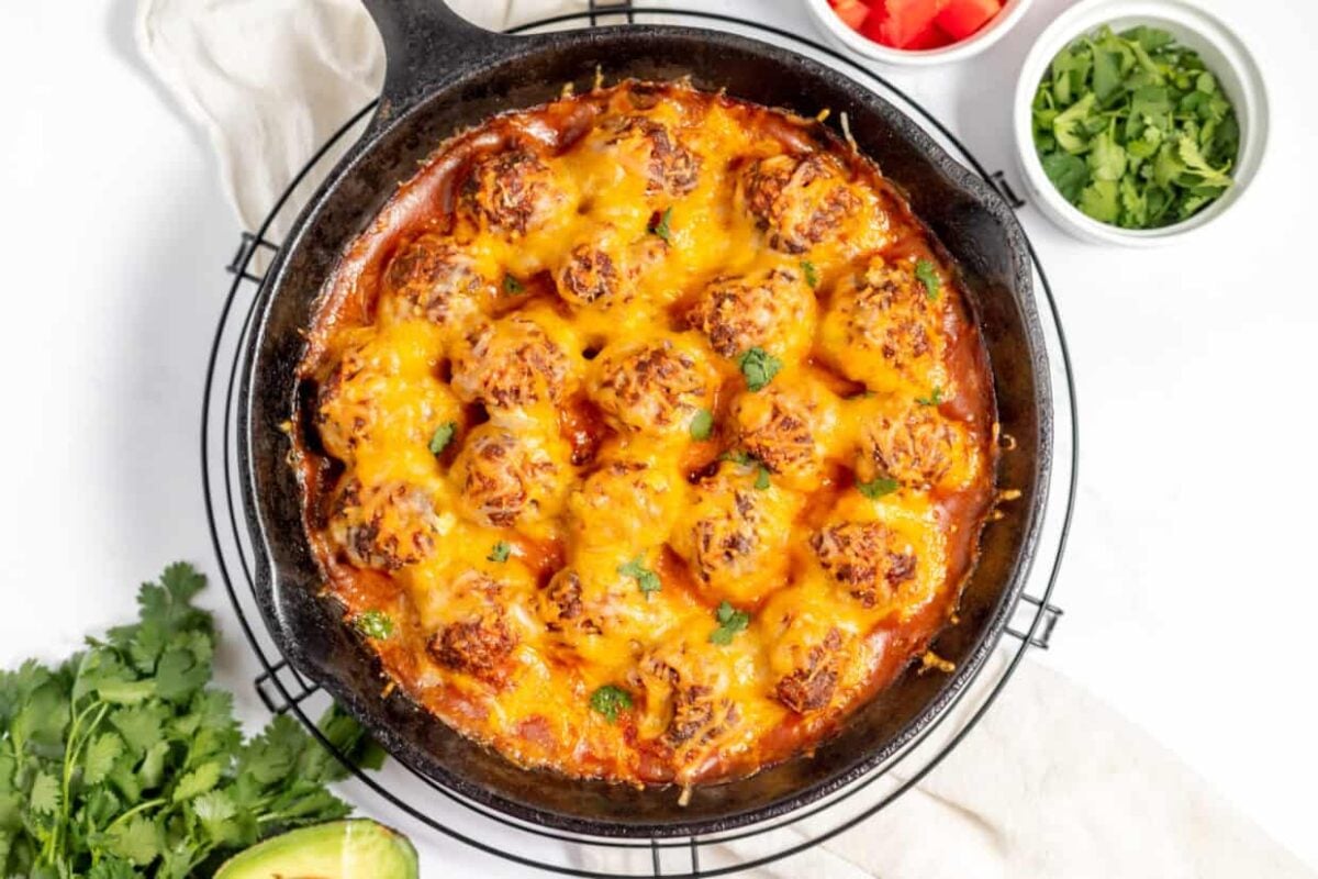 A cast iron skillet filled with baked meatballs topped with melted cheese, garnished with fresh cilantro—like a twist on cheesy enchiladas—with small bowls of chopped tomatoes and cilantro nearby.