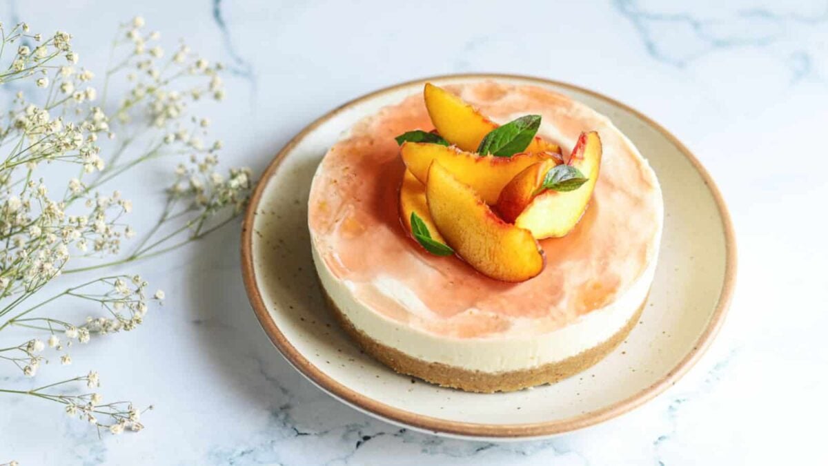 A round cheesecake with a light texture and a graham cracker crust, topped with peach slices and basil leaves, rests on a beige plate against a marble background—a perfect choice for gluten free desserts lovers.