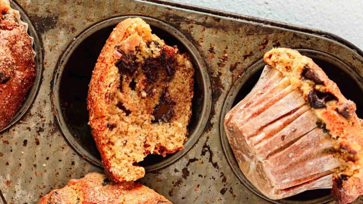 Chocolate chip muffins in a baking tray, one muffin cut in half revealing sweet layers of chocolate pieces inside, with another muffin still in a paper liner and boasting a light texture.