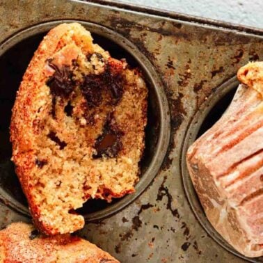 Chocolate chip muffins in a baking tray, one muffin cut in half revealing sweet layers of chocolate pieces inside, with another muffin still in a paper liner and boasting a light texture.