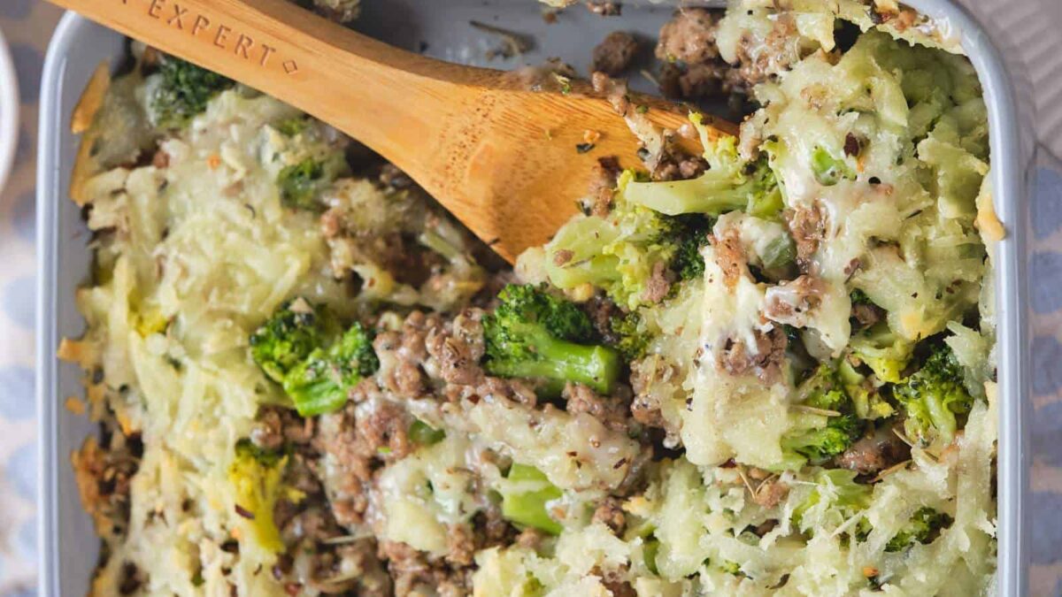 A close-up of a casserole dish containing a baked mixture of ground meat, broccoli, and shredded cheese, with a wooden spoon partially serving a portion—perfect for those seeking flavorful recipes or hearty broccoli dinner recipes.