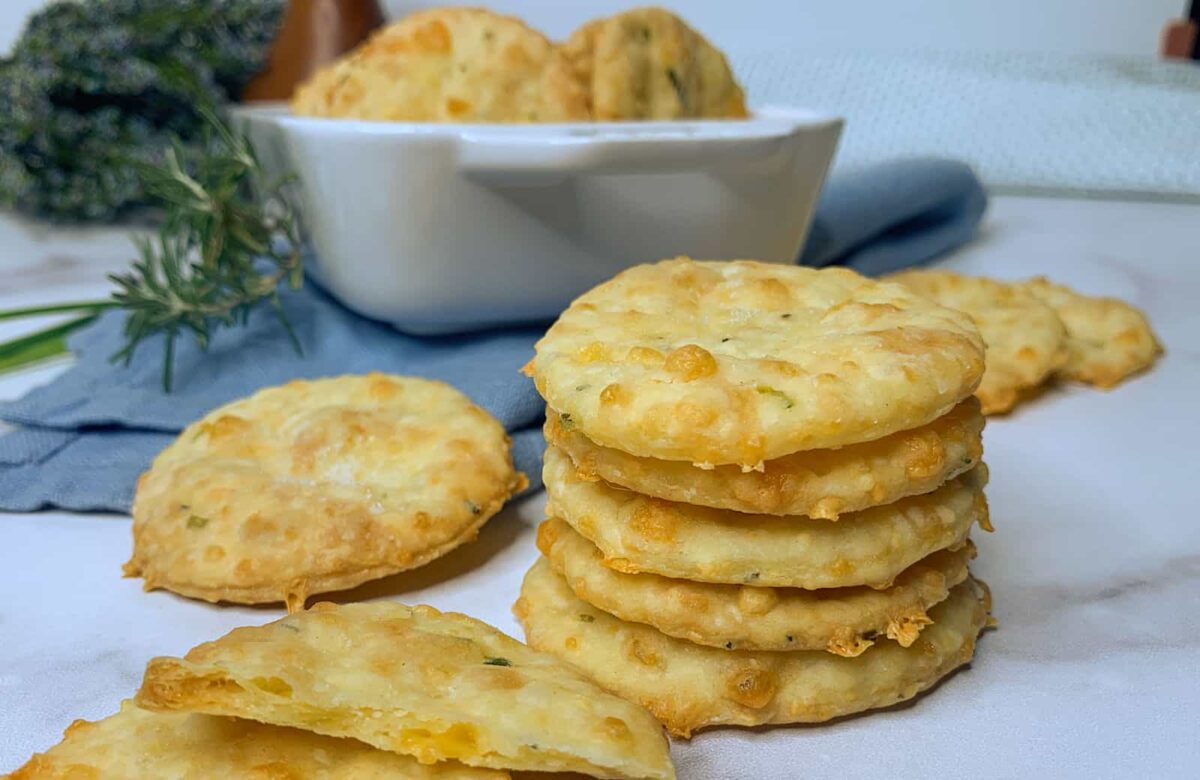 A stack of round, golden-brown crackers with herbs sits on a white surface, offering a crunchy and salty bite, with more crackers and snack chips in a bowl in the background.