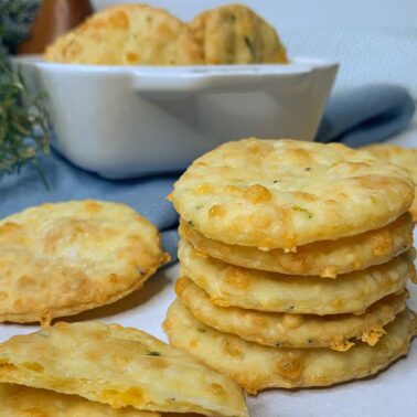 A stack of round, golden-brown crackers with herbs sits on a white surface, offering a crunchy and salty bite, with more crackers and snack chips in a bowl in the background.