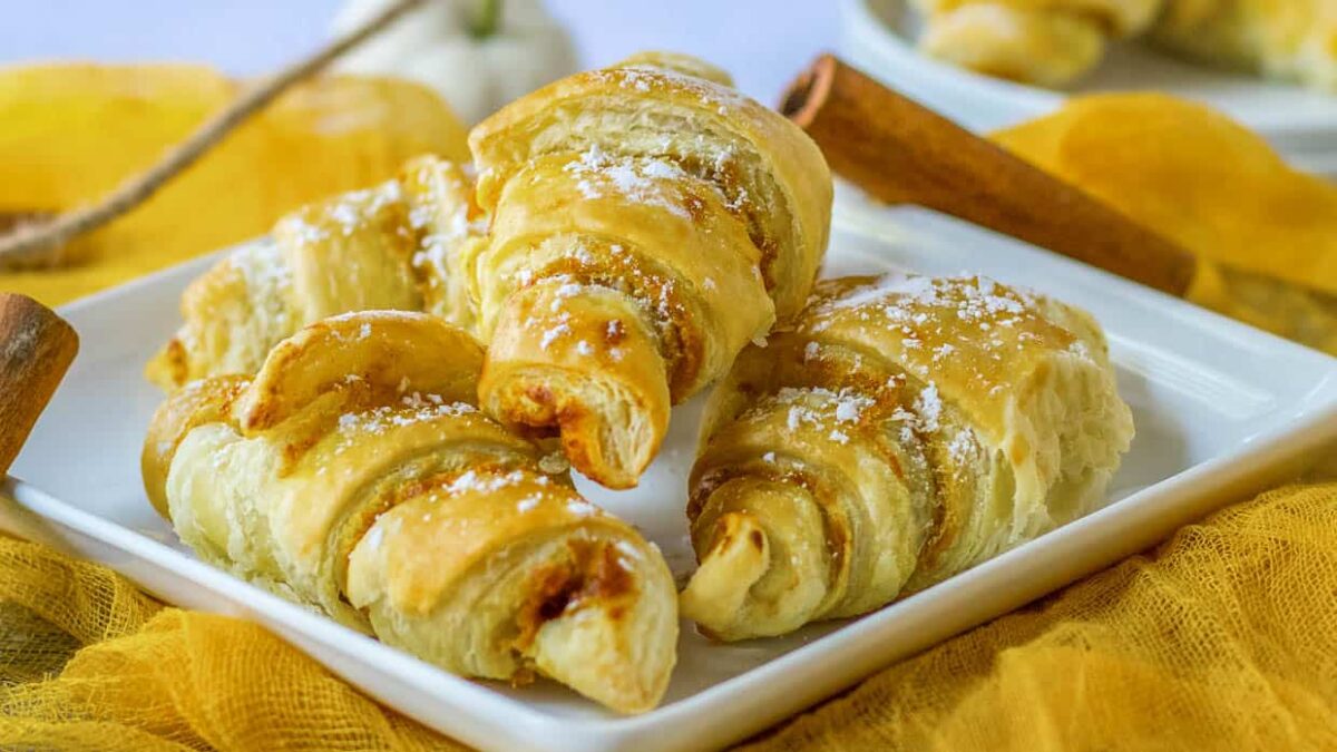 Four golden-brown croissants, sweet delights with a dusting of powdered sugar, are arranged on a white rectangular plate.