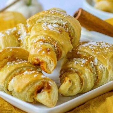 Four golden-brown croissants, sweet delights with a dusting of powdered sugar, are arranged on a white rectangular plate.