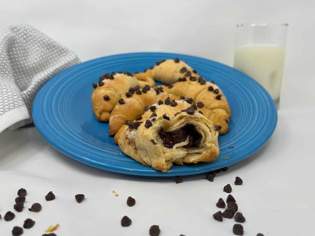 A blue plate with chocolate chip-filled croissants, one cut open to show the sweet delights inside. Scattered chocolate chips and a glass of milk are in the background.