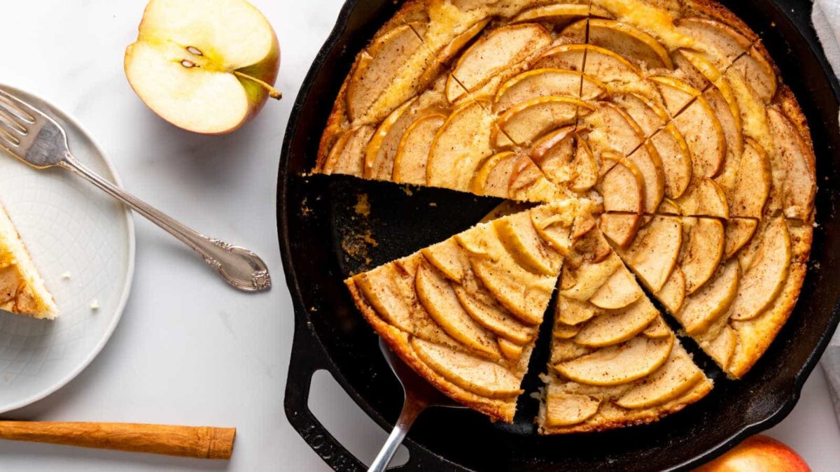 A sliced apple tart in a cast iron skillet, with a serving spatula and a slice on a plate nearby; half an apple and utensils are also visible—perfect for those seeking nut-free dessert recipes.