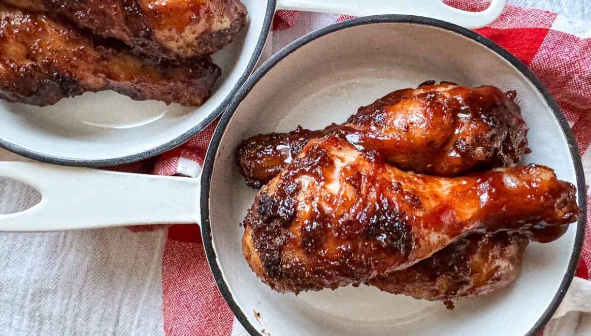 Two baked barbecue chicken drumsticks with charred edges are served in a white dish, with more chicken pieces in a similar dish nearby on a red and white cloth—a perfect fit for lovers of bold sauces and Smoky BBQ recipes.