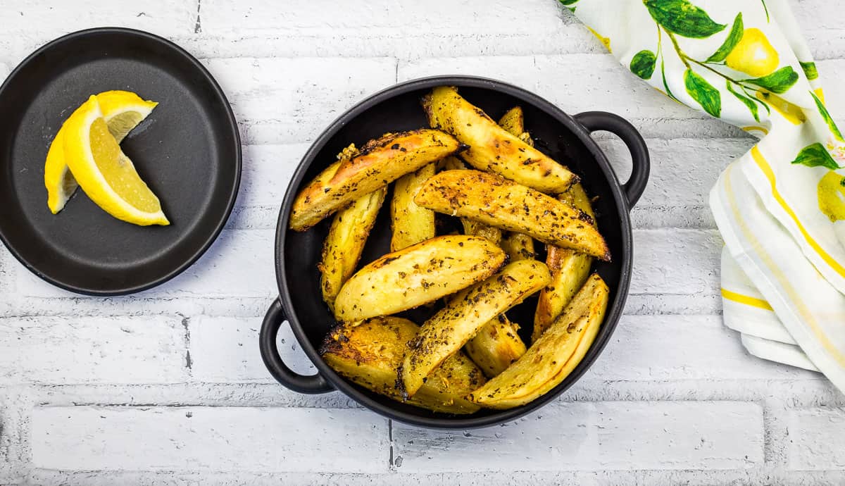 Black bowl filled with seasoned potato wedges, one of the ultimate comfort food side dishes, on a white brick surface, with a small plate holding two lemon wedges nearby.