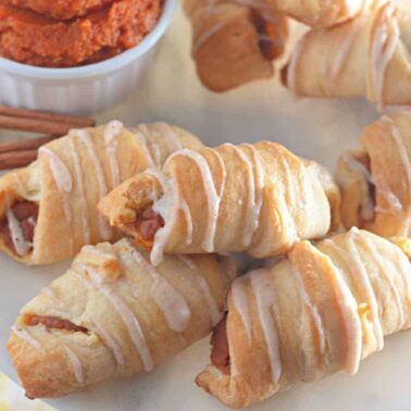 A plate of crescent rolls drizzled with icing, served with a small bowl of orange-colored dip and cinnamon sticksโperfect for those seeking safe, nut-free desserts.