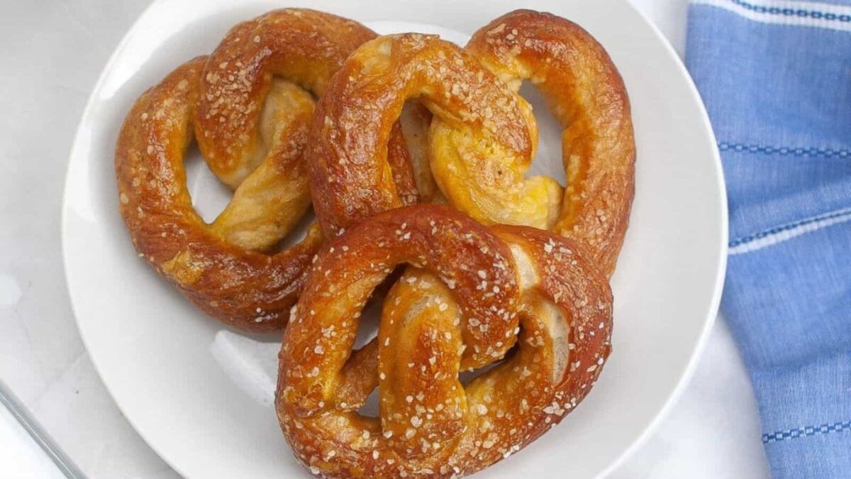 Three sourdough soft pretzels are arranged on a white plate next to a blue and white striped napkin, bringing a cozy kitchen feel to the scene.