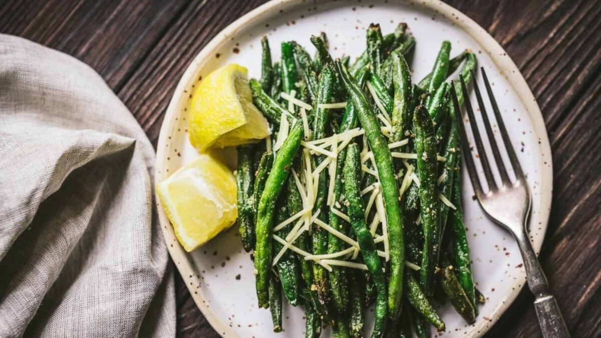 A plate of flavorful green beans topped with shredded cheese, served as bite-sized vegetarian appetizers alongside two lemon wedges and a fork.