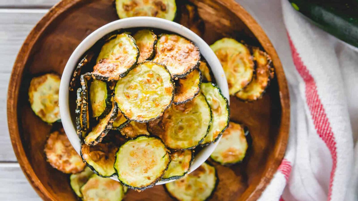 A bowl of bite-sized baked zucchini chips sits in a larger wooden bowl filled with more fresh vegetarian appetizers, next to a red-and-white striped napkin.