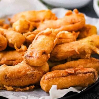 A plate of golden-brown fried banana fritters, perfect as bite-sized appetizers, served on parchment paper with a bowl of green chutney in the background.