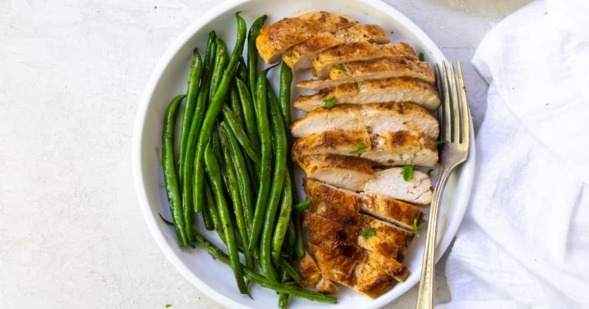 A white plate with sliced, sear-perfect grilled chicken breast and steamed green beans, accompanied by a fork on the side.