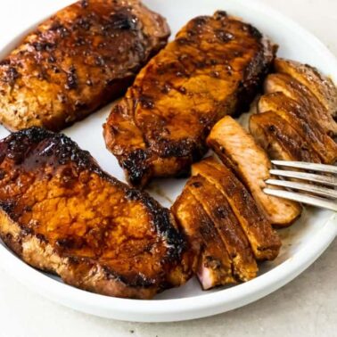 Three sear-perfect grilled pork chops, one sliced, served on a white plate with a fork on the sideโa delicious example of outdoor cooking.