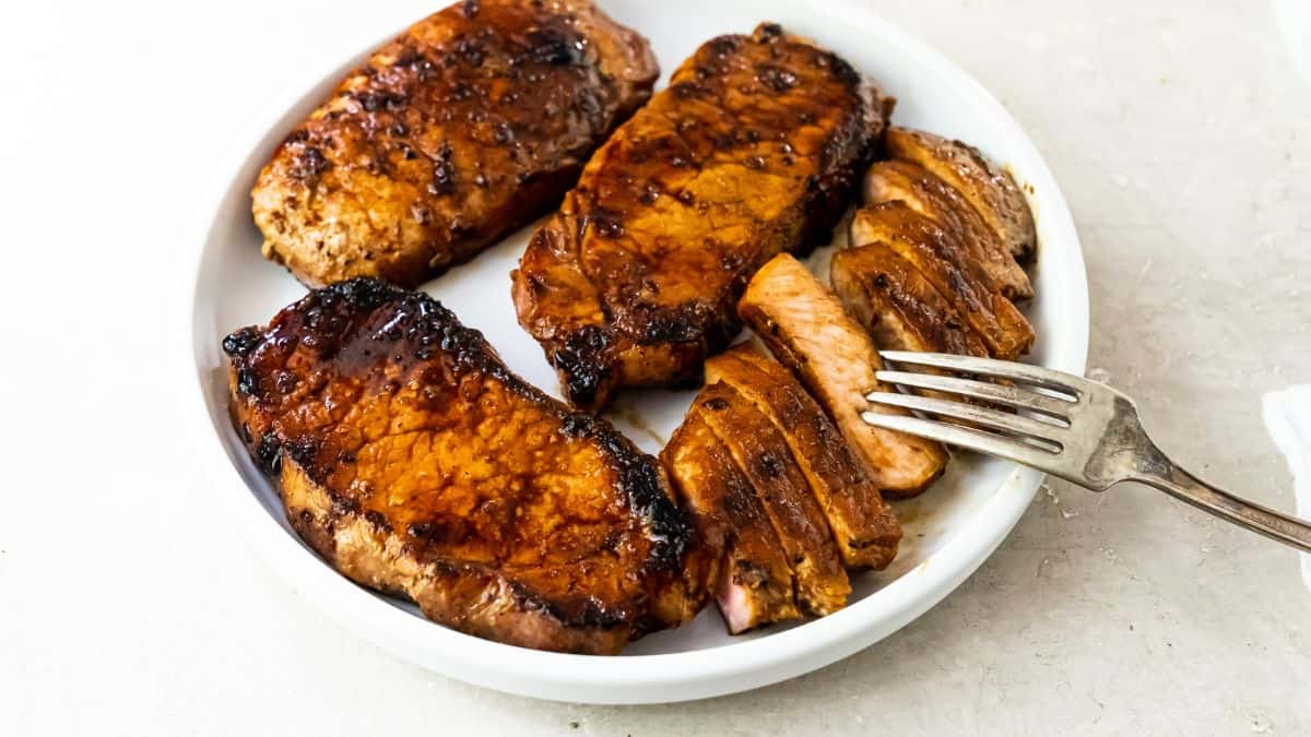 Three sear-perfect grilled pork chops, one sliced, served on a white plate with a fork on the side—a delicious example of outdoor cooking.