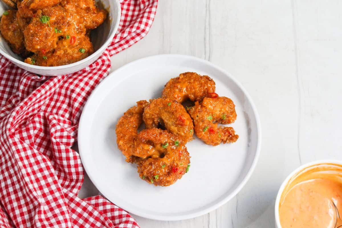 A white plate with four breaded and sauced shrimp, a bowl of shrimp, a red checkered cloth, and a bowl of dipping sauce on a white surface—perfect for Asian dinner recipes featuring bold spices and balanced flavors.