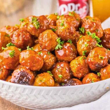 A white bowl filled with glazed meatballs featuring charred edges and garnished with chopped parsley sits on a wooden surface next to a drink can and a glass.