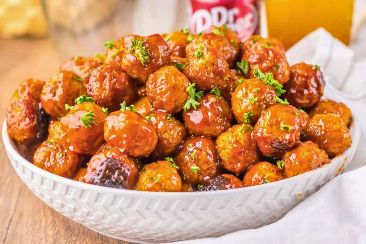 A white bowl filled with glazed meatballs featuring charred edges and garnished with chopped parsley sits on a wooden surface next to a drink can and a glass.
