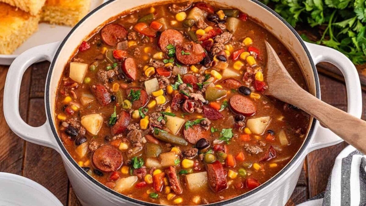 A pot of hearty stew containing sausage, beans, corn, potatoes, tomatoes, and vegetables boasts a rich flavor, with a wooden spoon resting inside.