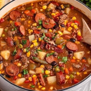 A pot of hearty stew containing sausage, beans, corn, potatoes, tomatoes, and vegetables boasts a rich flavor, with a wooden spoon resting inside.