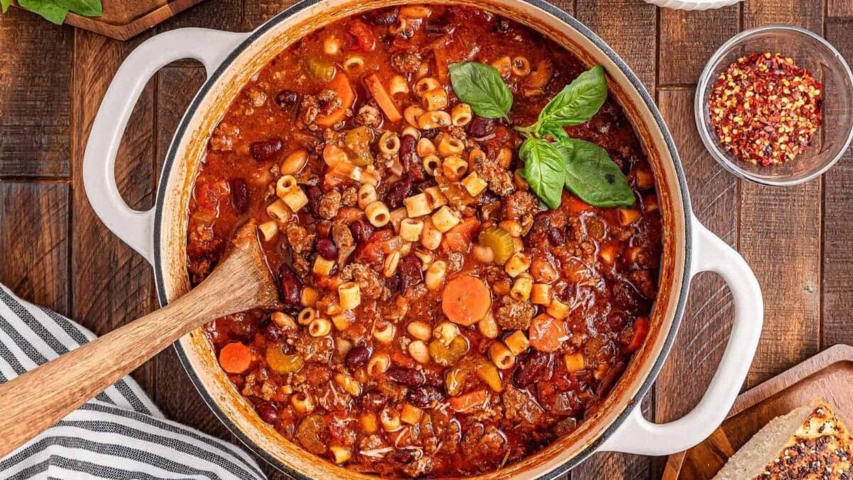 A large pot of hearty minestrone soup with rich flavor—packed with pasta, beans, vegetables, and ground meat—is garnished with fresh basil and sits on a wooden table with a wooden spoon inside. A perfect one pot meal!.