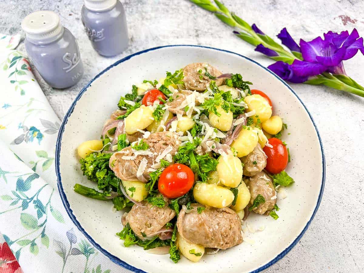 A bowl of skillet gnocchi with sausage, cherry tomatoes, broccoli rabe, and grated cheese, garnished with parsley. Salt and pepper shakers and a purple flower are nearby—perfect inspiration for your favorite gnocchi recipes.