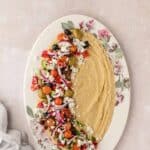 Oval plate with hummus spread on one side and a chopped Greek salad with olives, tomatoes, onion, and feta cheese on the other side, set on a light surface.