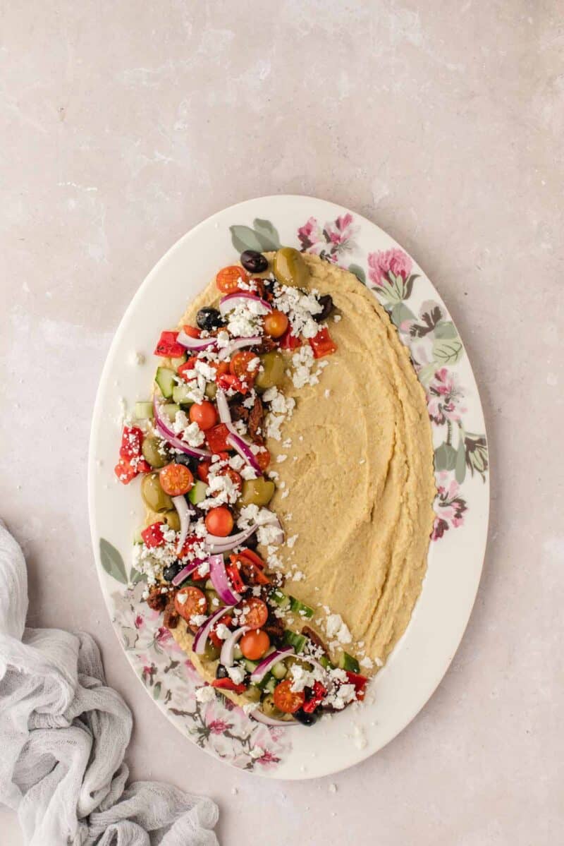 Oval plate with hummus spread on one side and a chopped Greek salad with olives, tomatoes, onion, and feta cheese on the other side, set on a light surface.